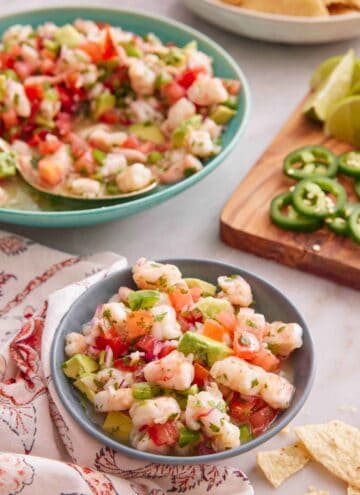 A bowl of shrimp ceviche with a platter in the back and cut garnishes on the side.