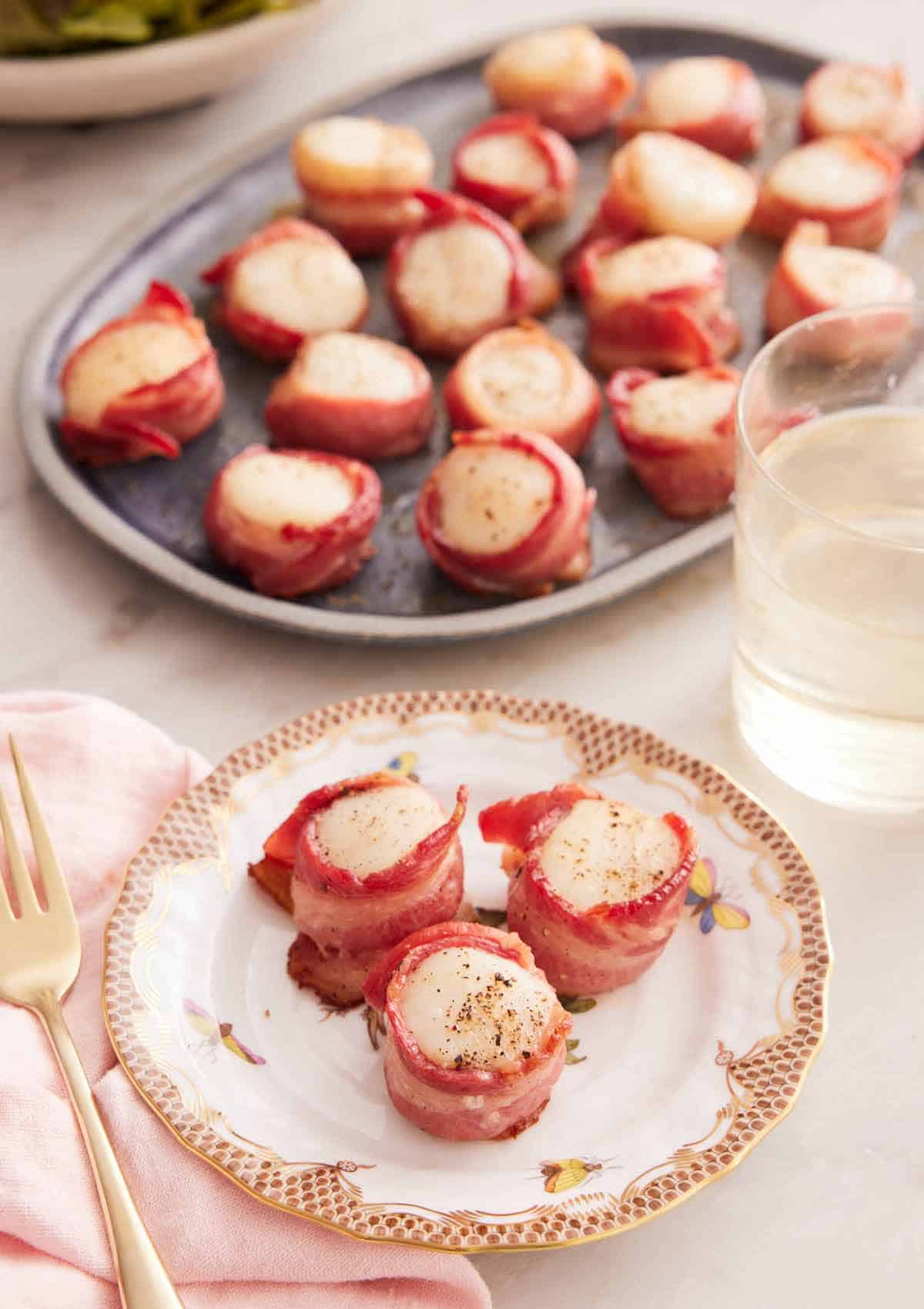 A plate with three bacon wrapped scallops with a platter in the back along with a glass of wine.