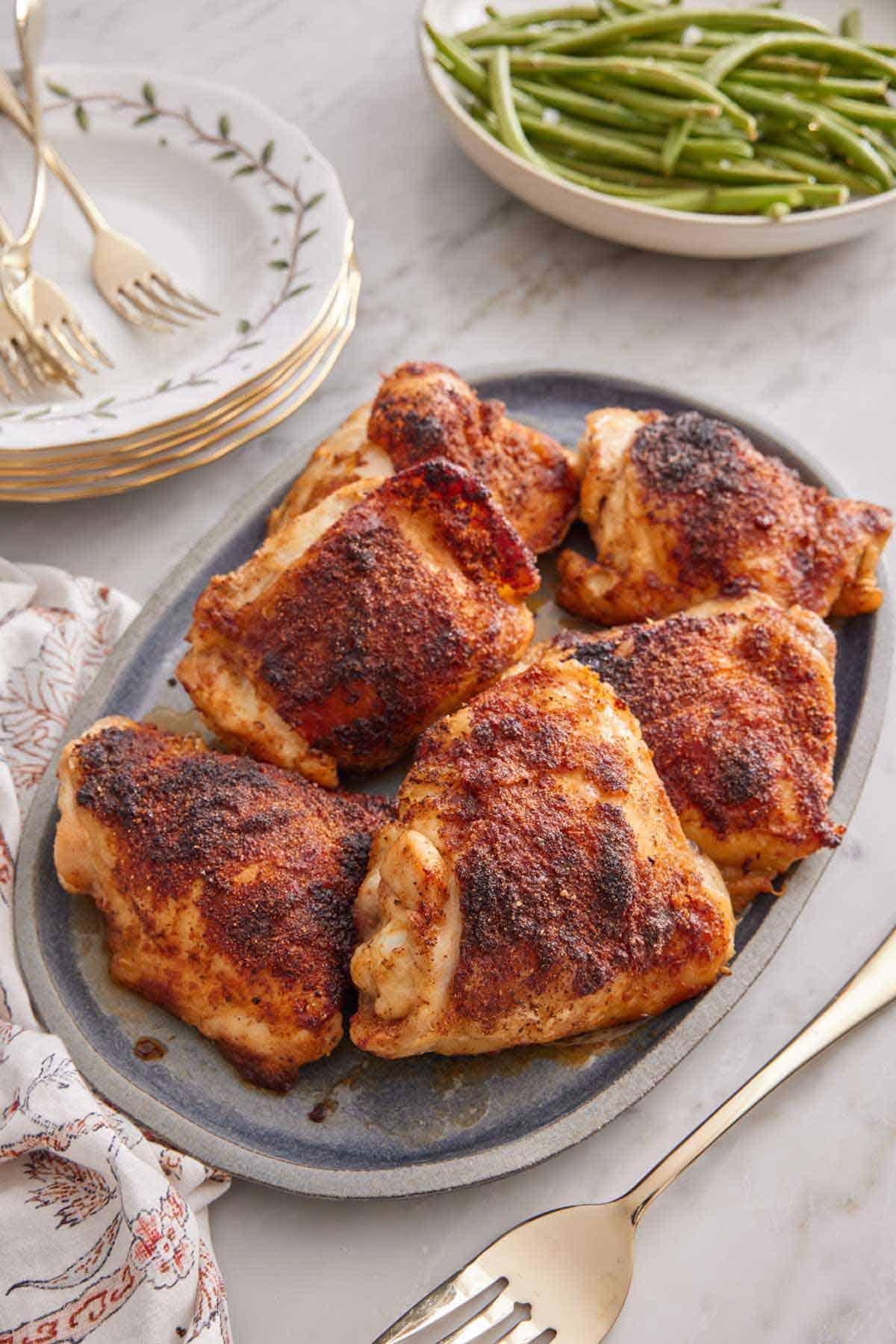 A platter of baked chicken thighs with a plate of green beans and stack of dishes with forks in the background.