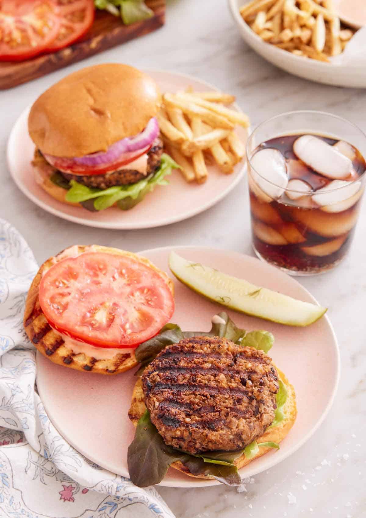 A plate with an opened veggie burger with a drink on the side with another veggie burger in the background with fries.