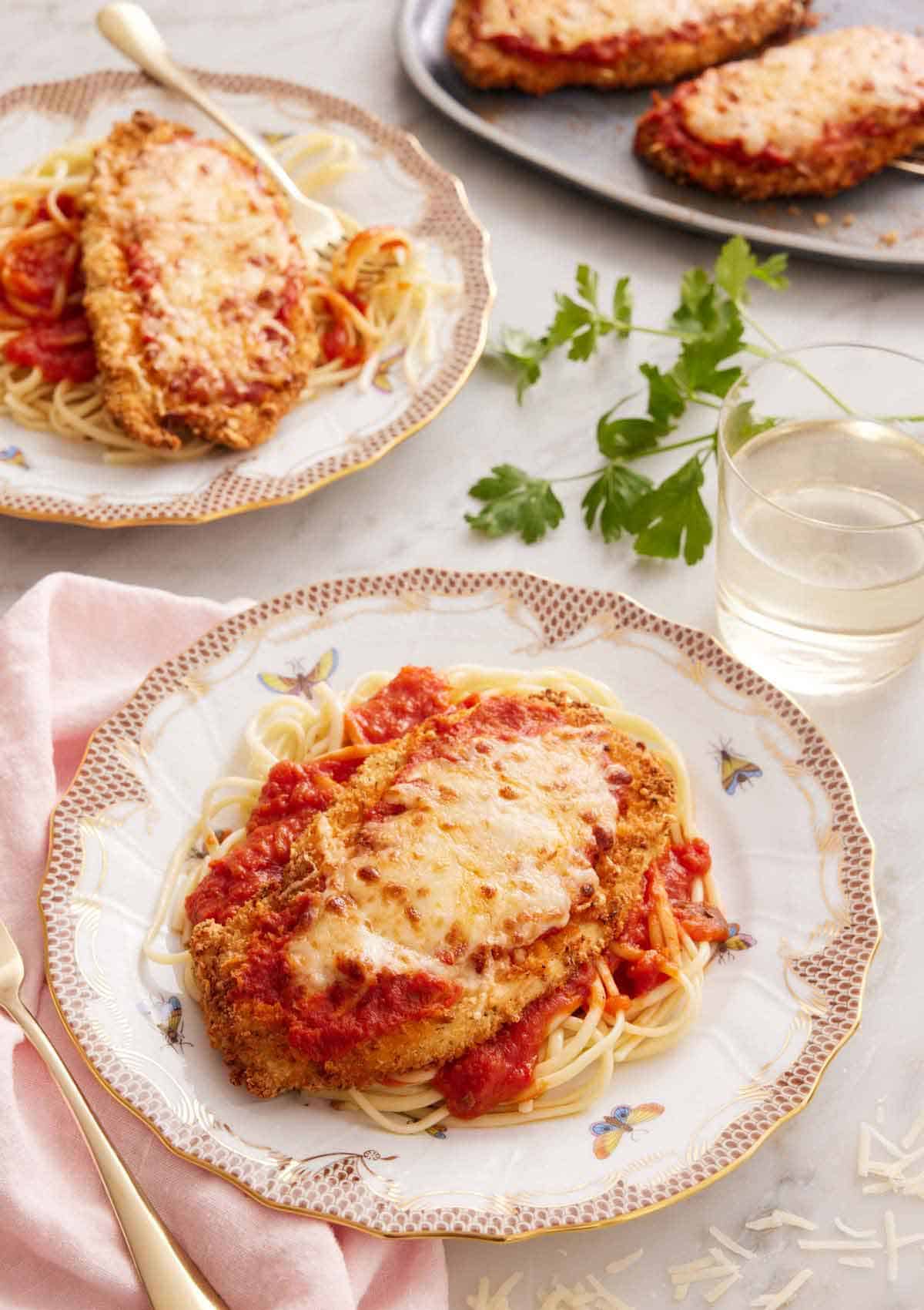 Two plates of air fryer chicken parmesan over spaghetti noodles with a glass of wine and parsley in between the plates.