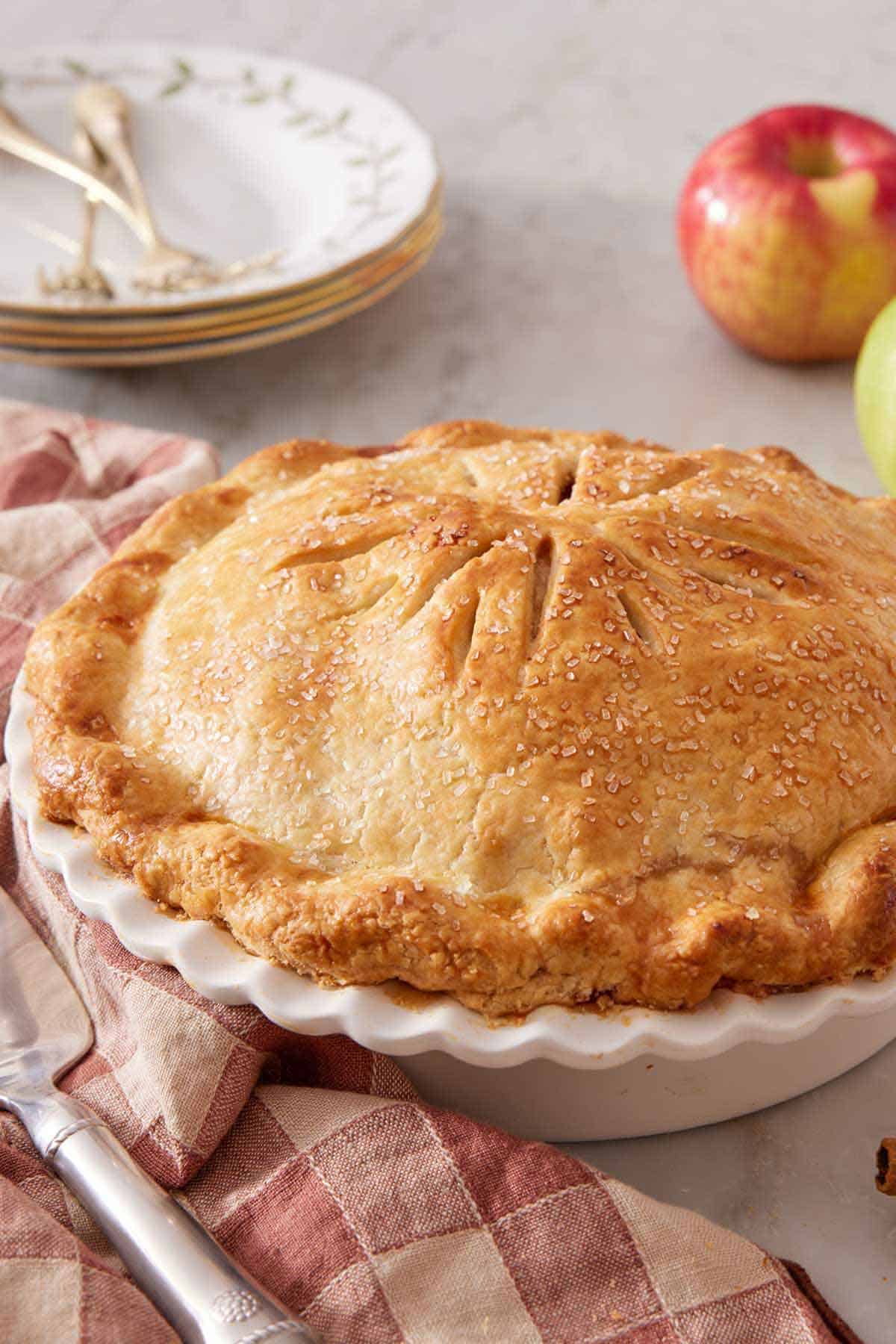 An apple pie in a white pie dish with a stack of plates and some apples in the background.