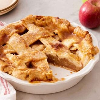 A white pie dish with an apple pie inside with a slice cut out. An apple and stack of plates in the back.