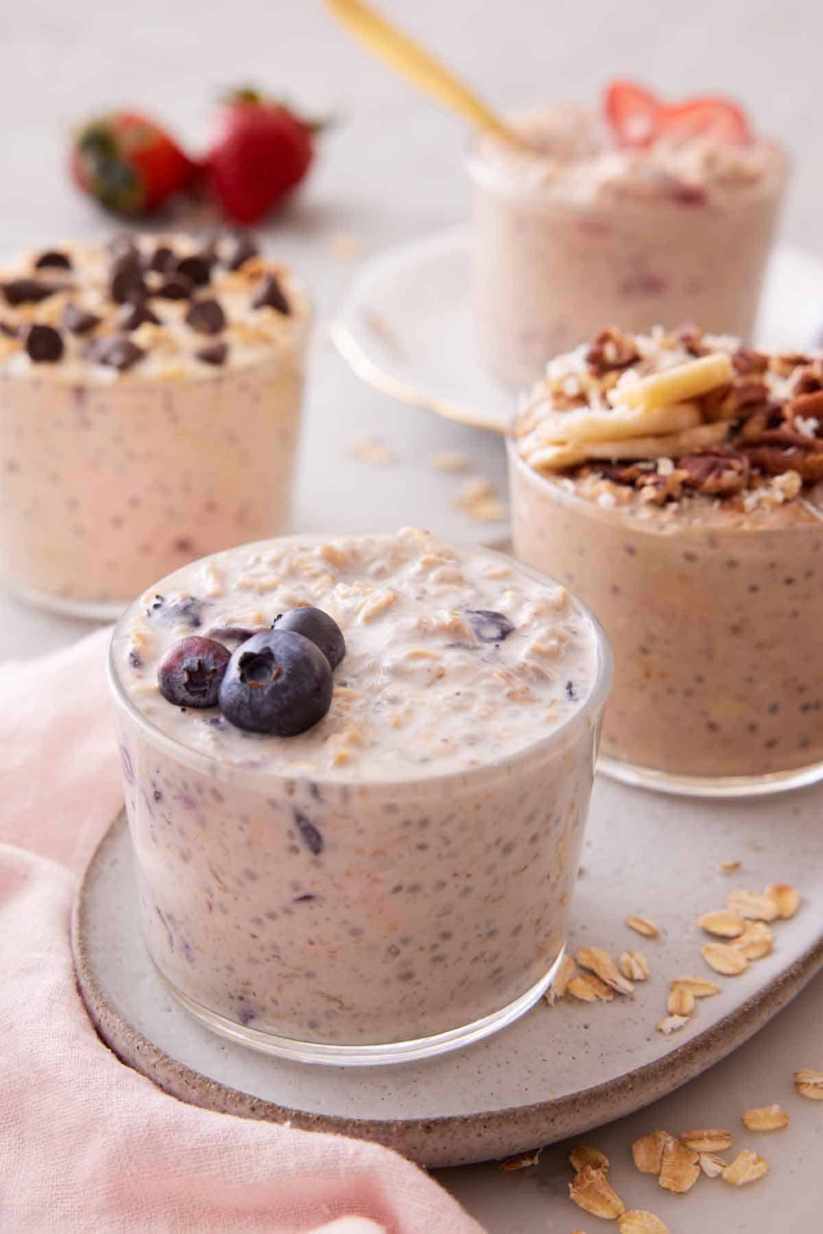 A platter with a glass of overnight oats topped with blueberries with additional glasses in the background with different toppings.