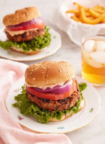 A plate with a black bean burger with a drink in the back along with a second burger and bowl of fries.