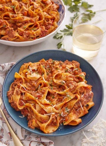 A bowl of bolognese with a glass of wine and a second bowl in the background.
