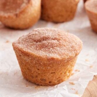 A cinnamon muffin on a wooden board lined with parchment with more muffins in the back.
