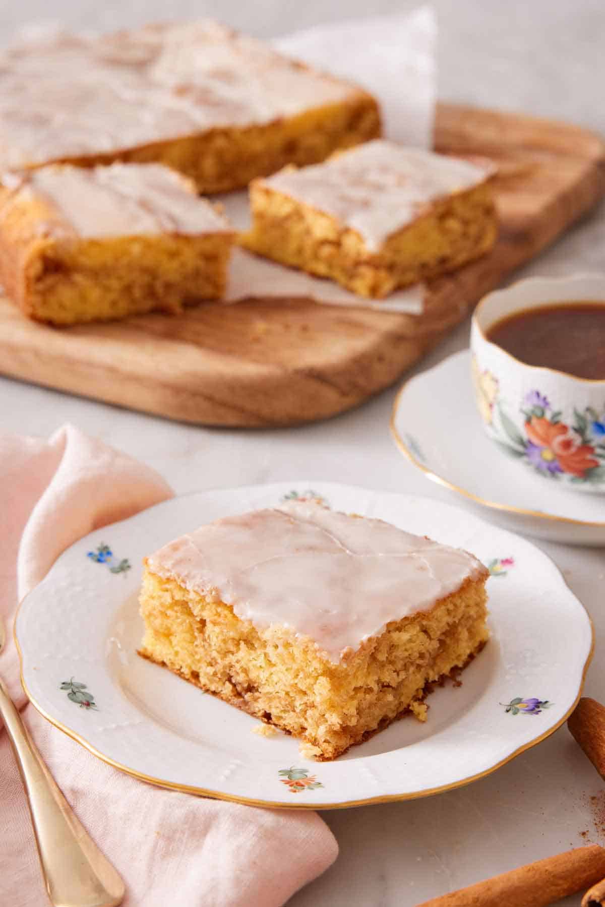 A slice of honey bun cake with a cup of coffee and the rest of the cake in the background.
