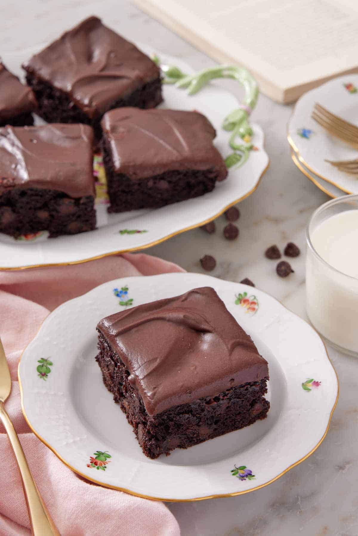 A plate with a slice of zucchini brownie with a platter in the background. Glass of milk on the side.