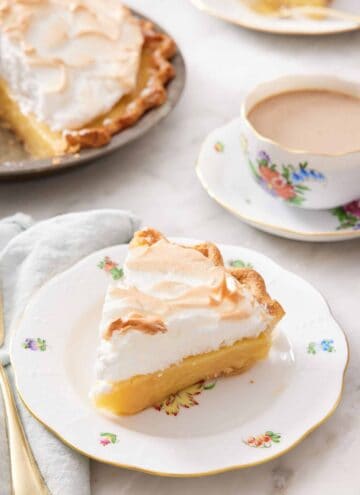 A plate with a slice of butterscotch pie with a cup of coffee in the background and the rest of the pie.