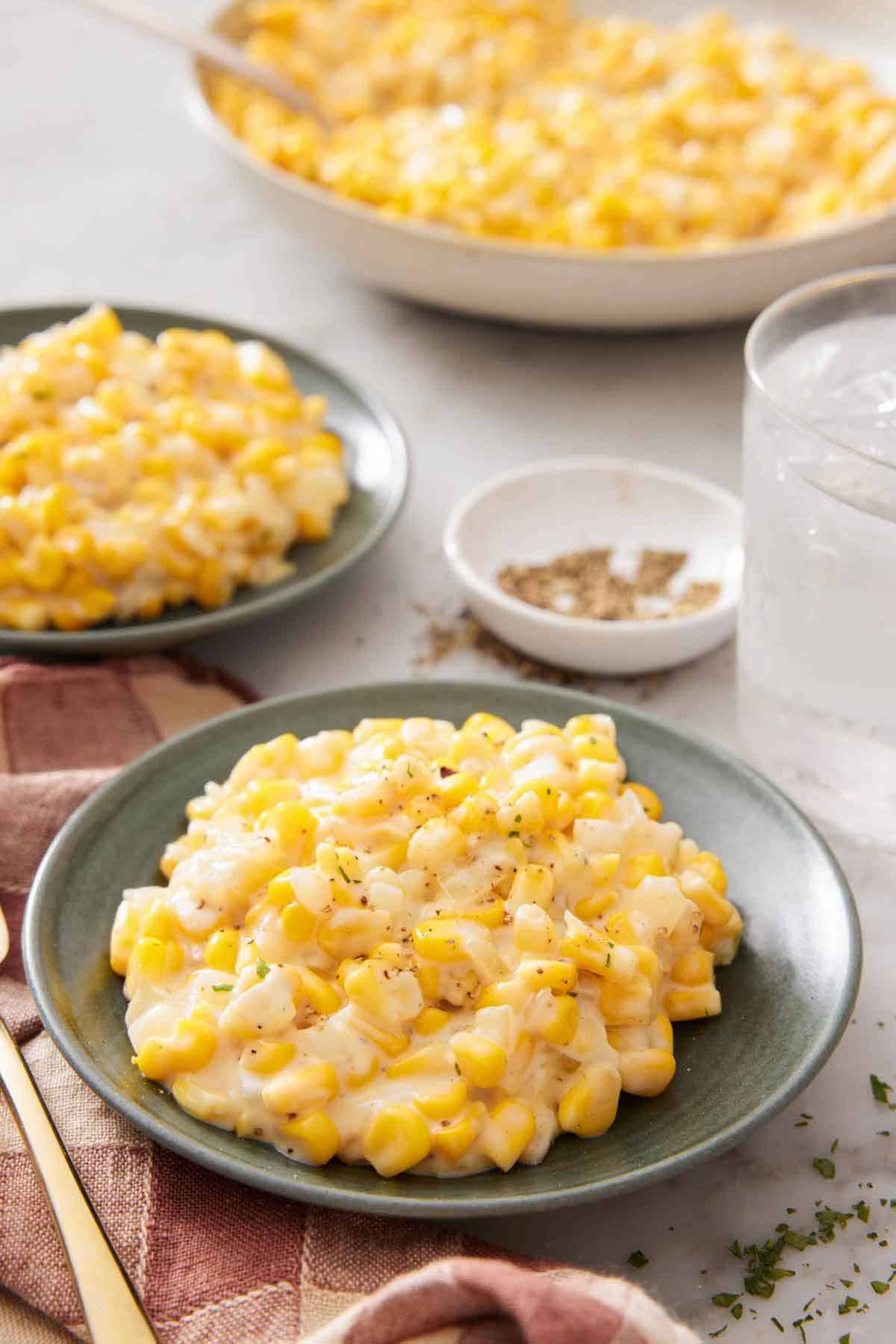 Two plates of creamed corn with a platter with more in the background along with a glass of water and bowl of pepper.