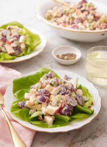 A plate with Waldorf salad on a lettuce leaf with another plated serving and a platter of salad in the background. A glass of wine and some pepper behind the plate.