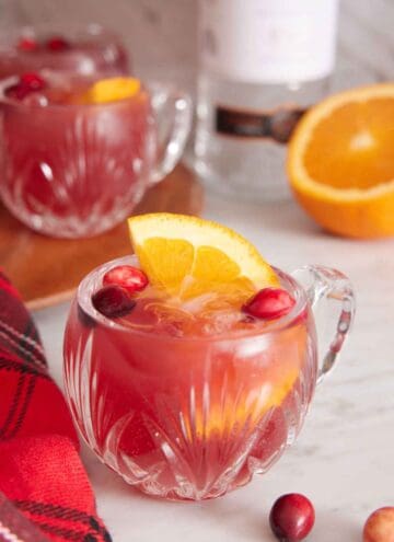 A glass of Christmas punch with orange and cranberries with more in the background along with a cut orange and bottle of vodka.