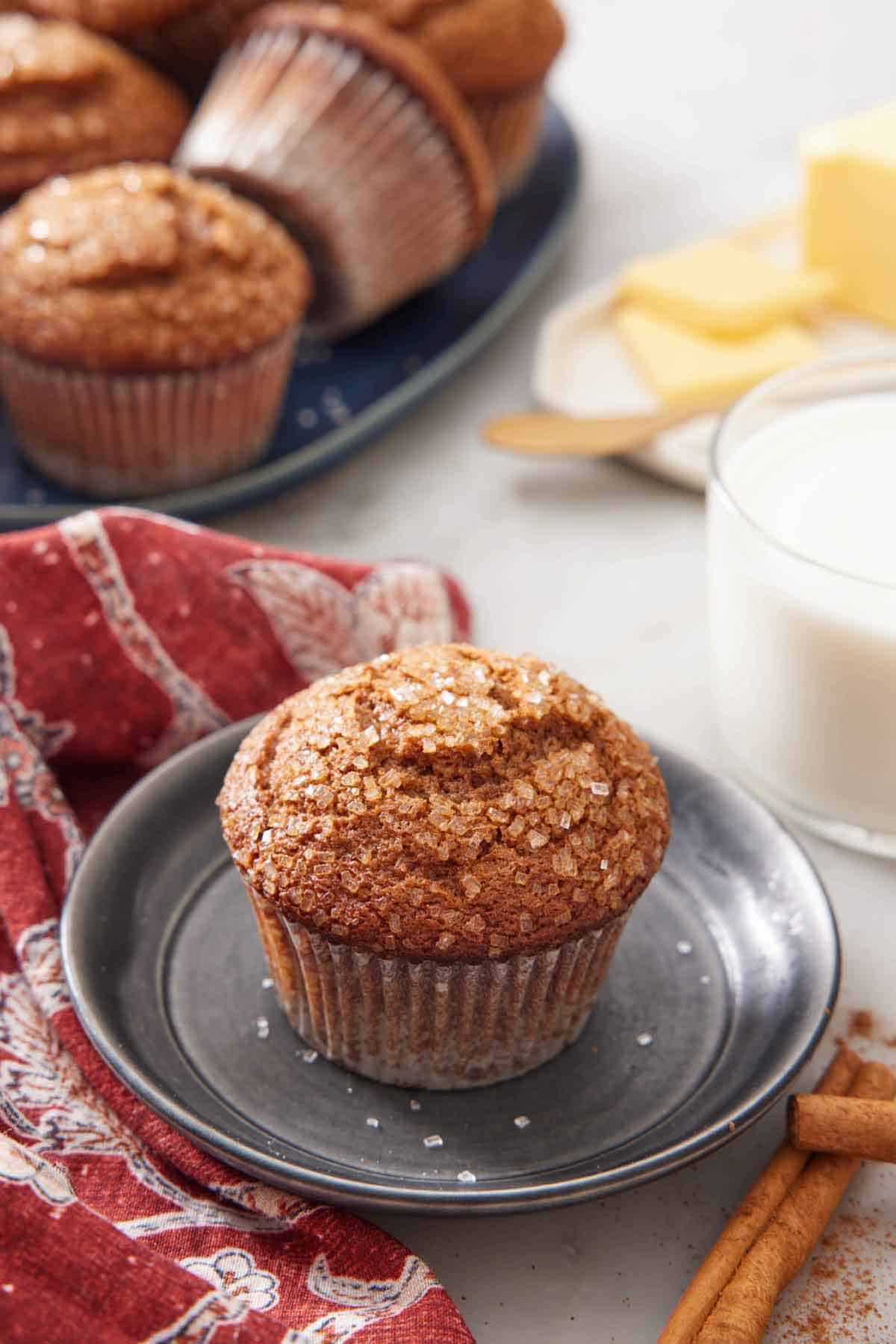 A plate with a gingerbread muffins with a glass of milk and platter of more muffins in the background.