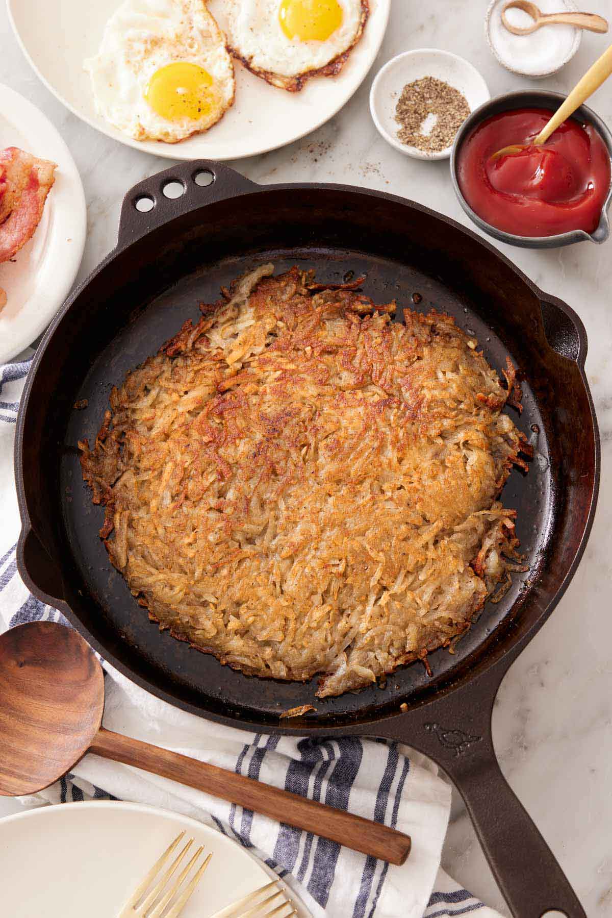 Overhead view of hash browns on a cast iron skillet. Some ketchup, salt, pepper, and fried eggs off to the side.