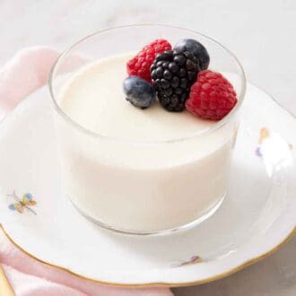 A glass with panna cotta topped with berries on a plate. A spoon beside it.