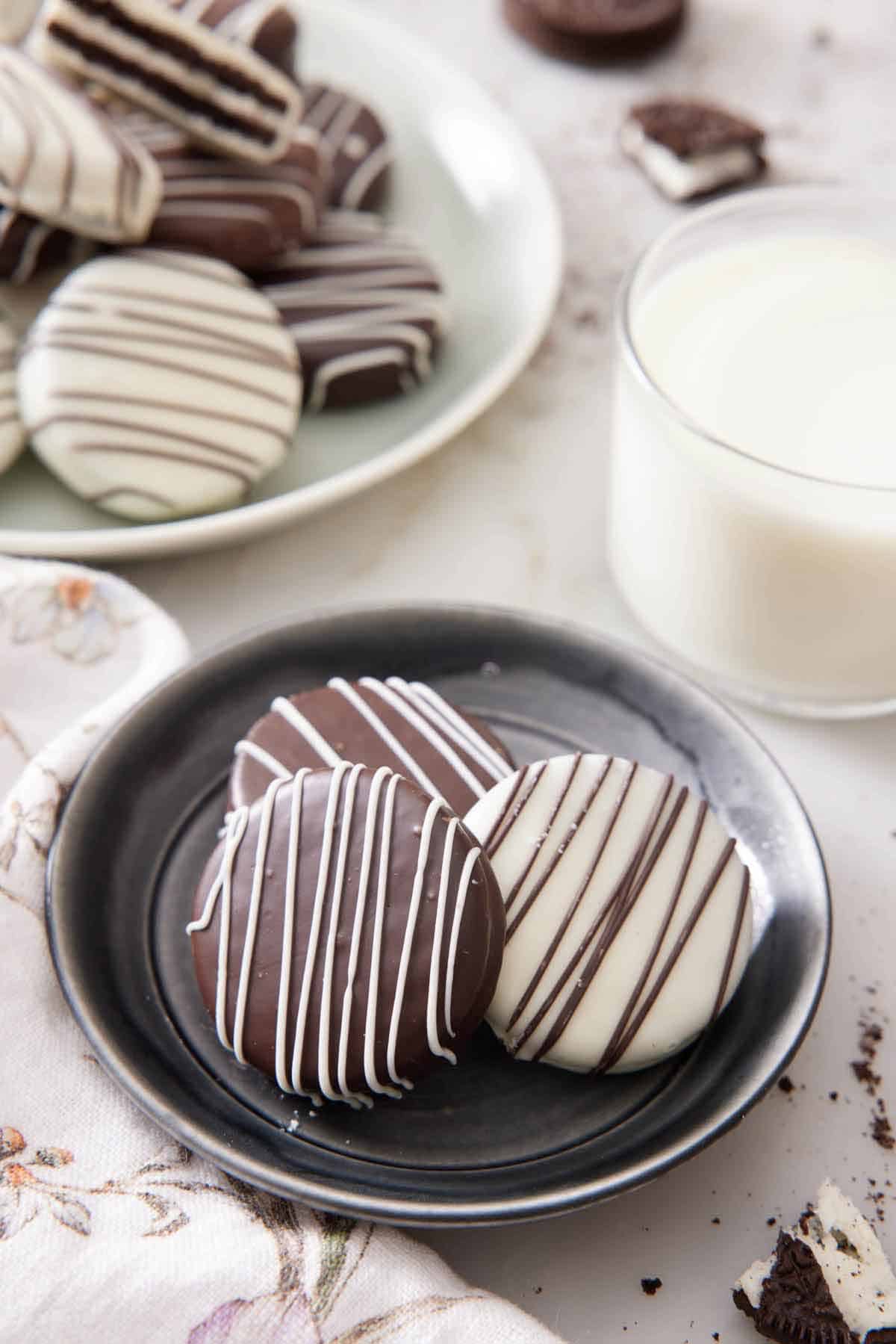 A plate with three chocolate covered oreos with a glass of milk and more oreos on a platter in the background.