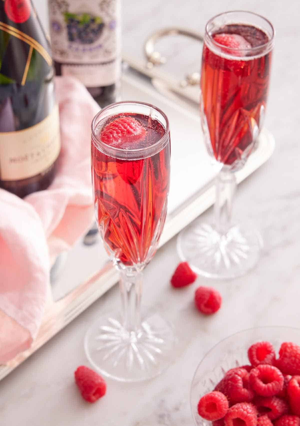 Two glasses of Kir Royale with a bowl of raspberries and the bottles of alcohol in the back.
