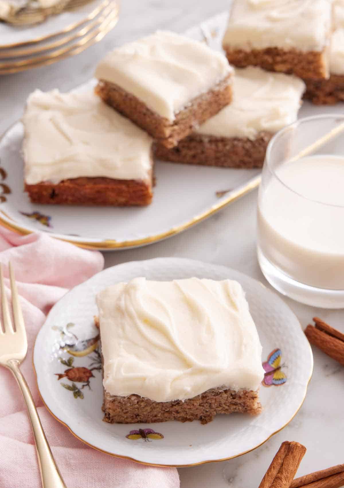A plate with a slice of banana bar with a glass of milk and platter of more bars in the background.