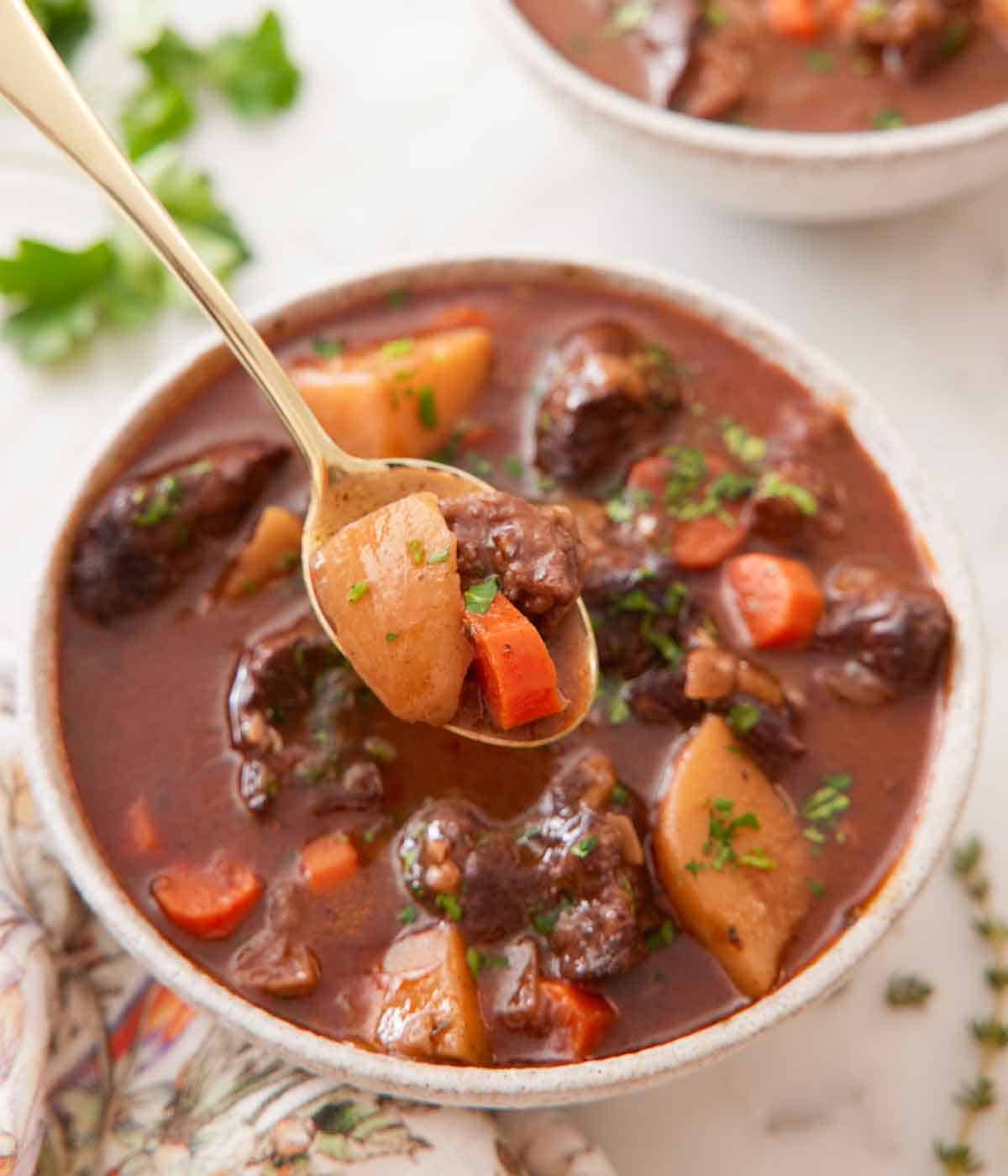 A spoonful of beef stew lifted up in front of a bowl of stew showing a potato, carrot, and piece of beef.