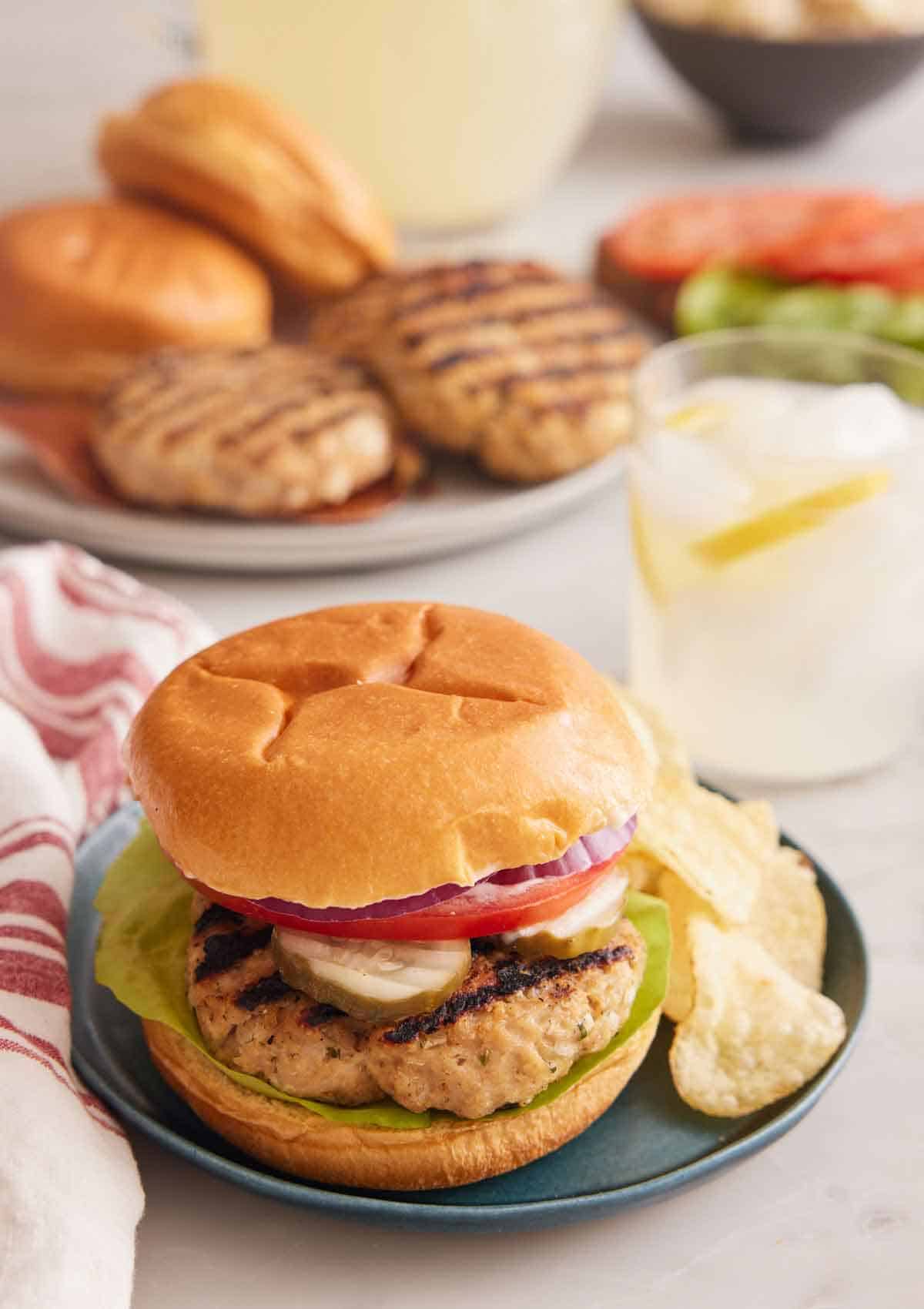 A plate with a chicken burger with potato chips with a drink in the background and burger components on plates.