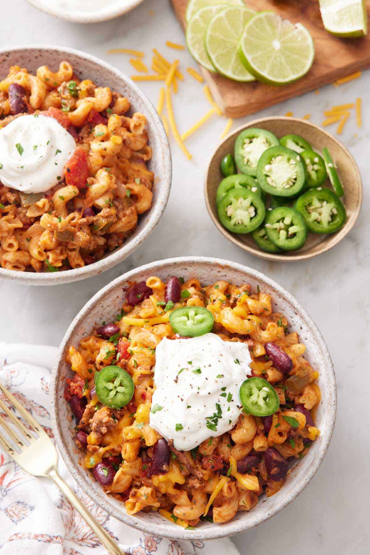 Two bowls of chili mac topped with sour cream with a bowl of sliced jalapeño and sliced limes.