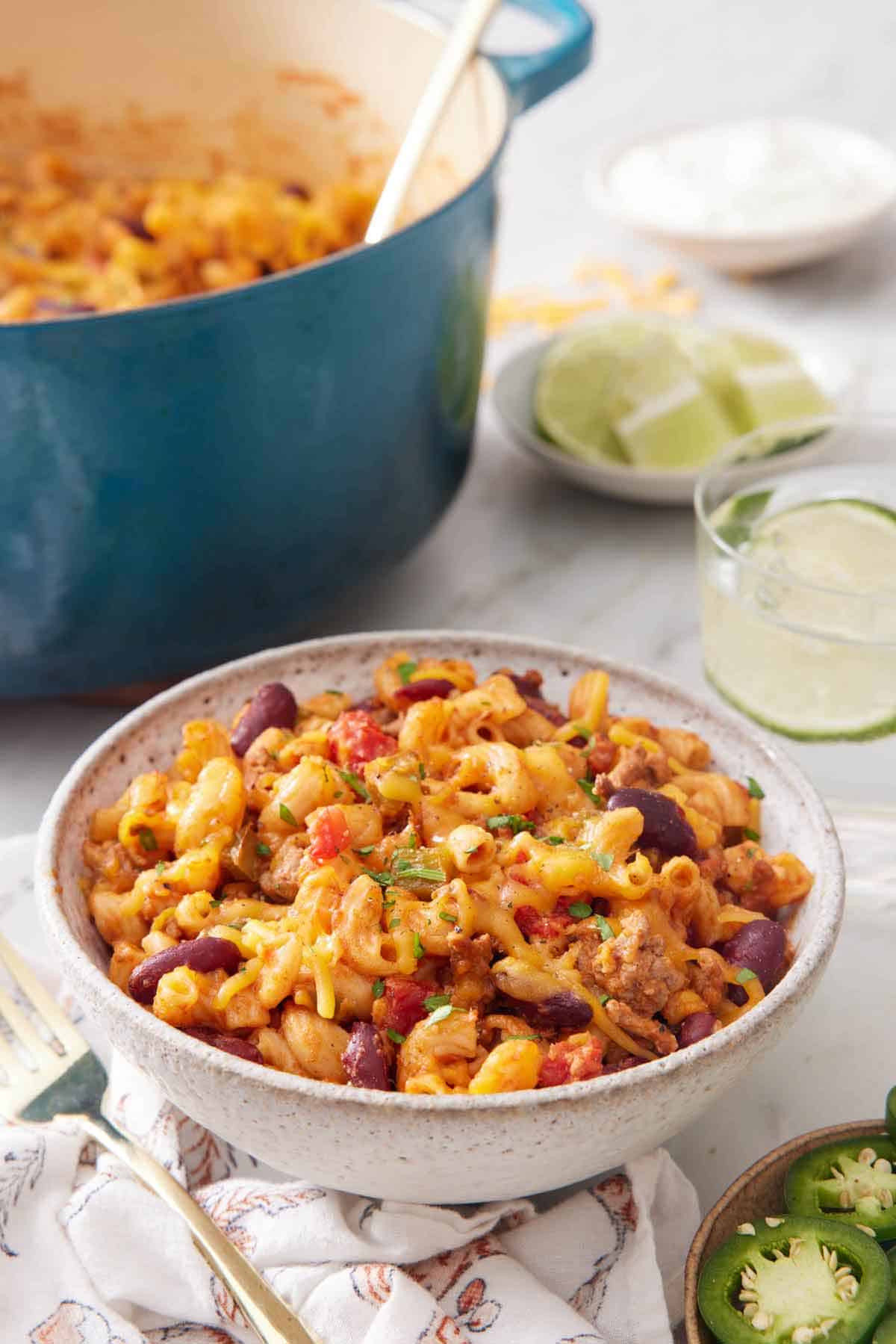 A bowl of chili mac with a drink in the background along with a bowl of limes and a pot of more chili mac.