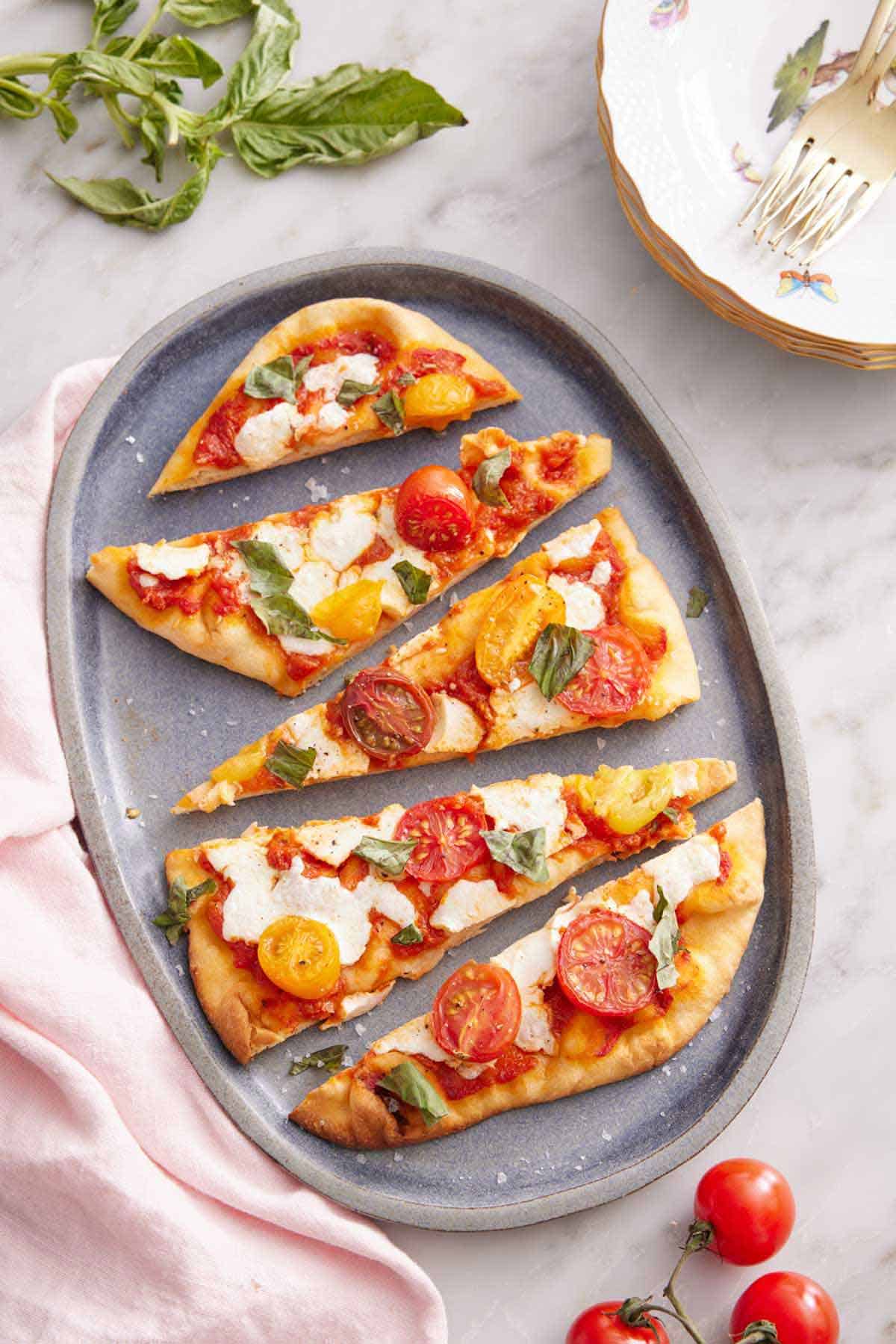 Overhead view of a platter of flatbread pizza sliced into triangle pieces. Basil, tomatoes, a stack of plates, and forks in the back.