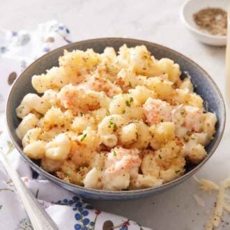 A bowl of lobster mac and cheese with a drink in the background along with a bowl of pepper with a napkin and fork beside it.