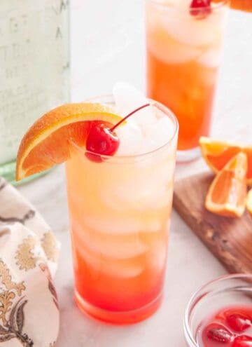 A glass of tequila sunrise with an orange slice and maraschino cherry as garnish. A second glass in the background along with the cut orange slices and tequila bottle.