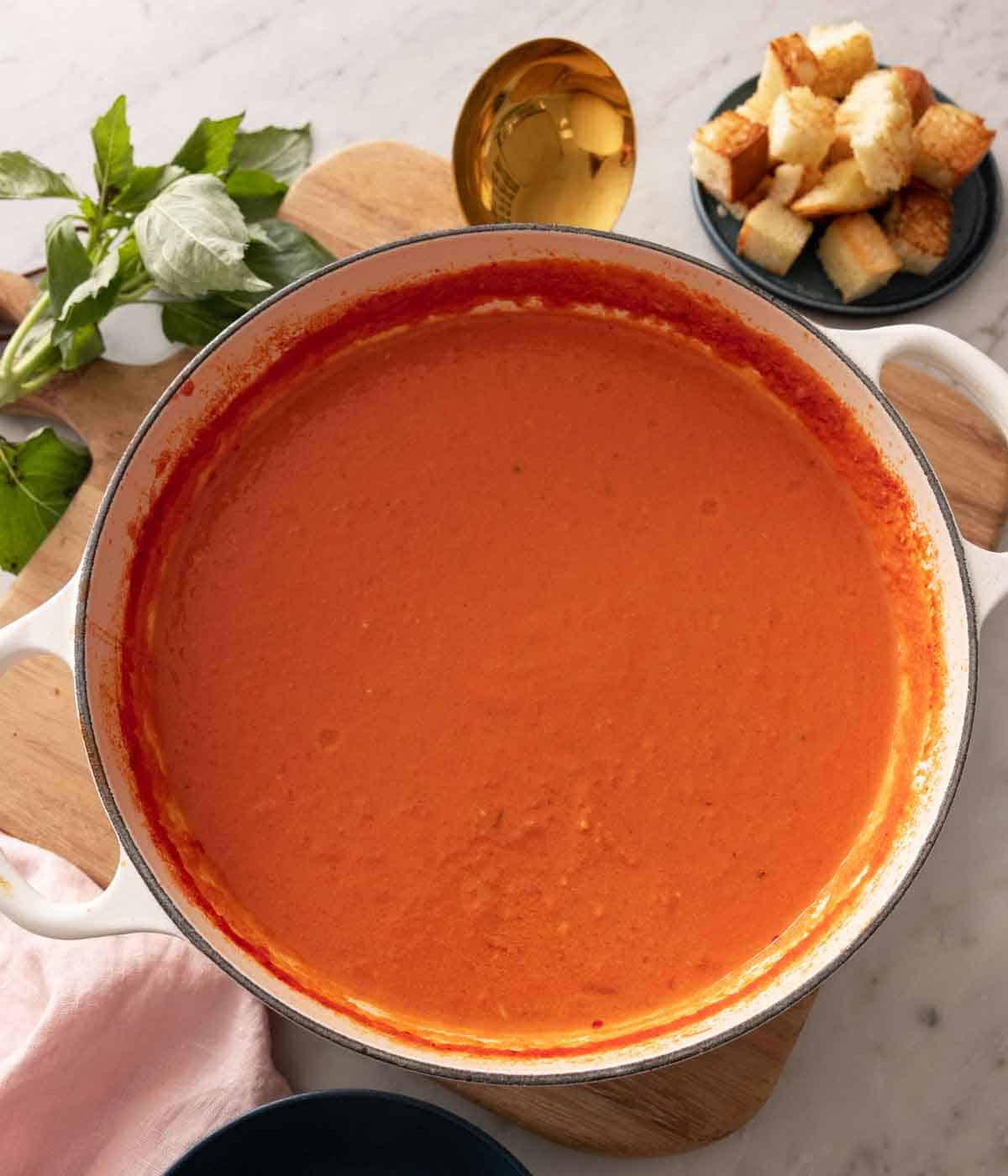 Overhead view of a white Dutch oven containing tomato soup. Basil and bread off to the side.