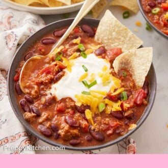 Pinterest graphic of a bowl of turkey chili topped with sour cream, cheese, green onions, and two chips with a spoon in the bowl.