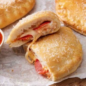 A calzone cut in half with one half stacked on top of the other. Two more in the background with a bowl of pizza sauce on the side.