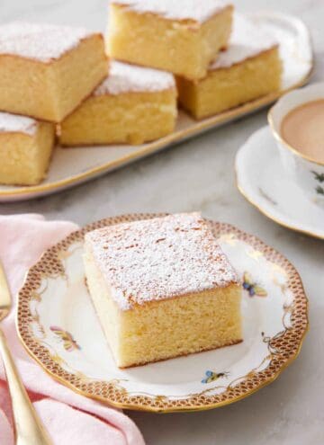 A slice of hot milk cake topped with a dusting of powdered sugar. A platter with more pieces in the back.