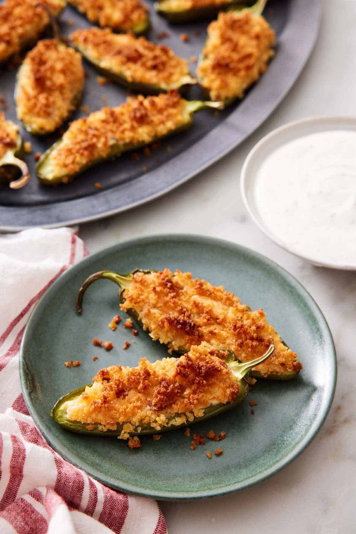 A plate with two jalapeno poppers with a bowl of dipping sauce on the side. A platter with more jalapeno poppers in the background.