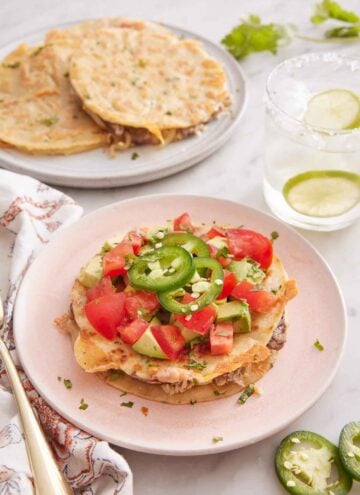 A plate of mulita topped with tomatoes, avocados, and jalapenos. A drink in the back and more mulitas on a plate.