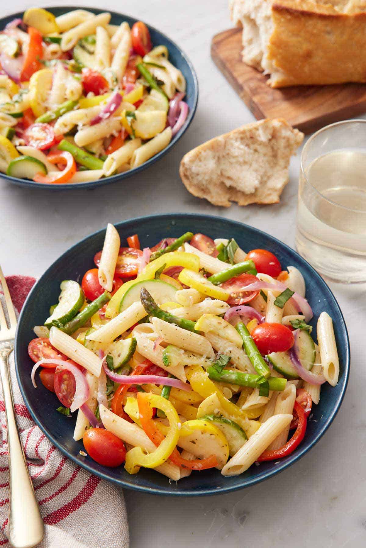 A bowl of pasta primavera with a second bowl in the background and bread torn on the side with a glass of wine.
