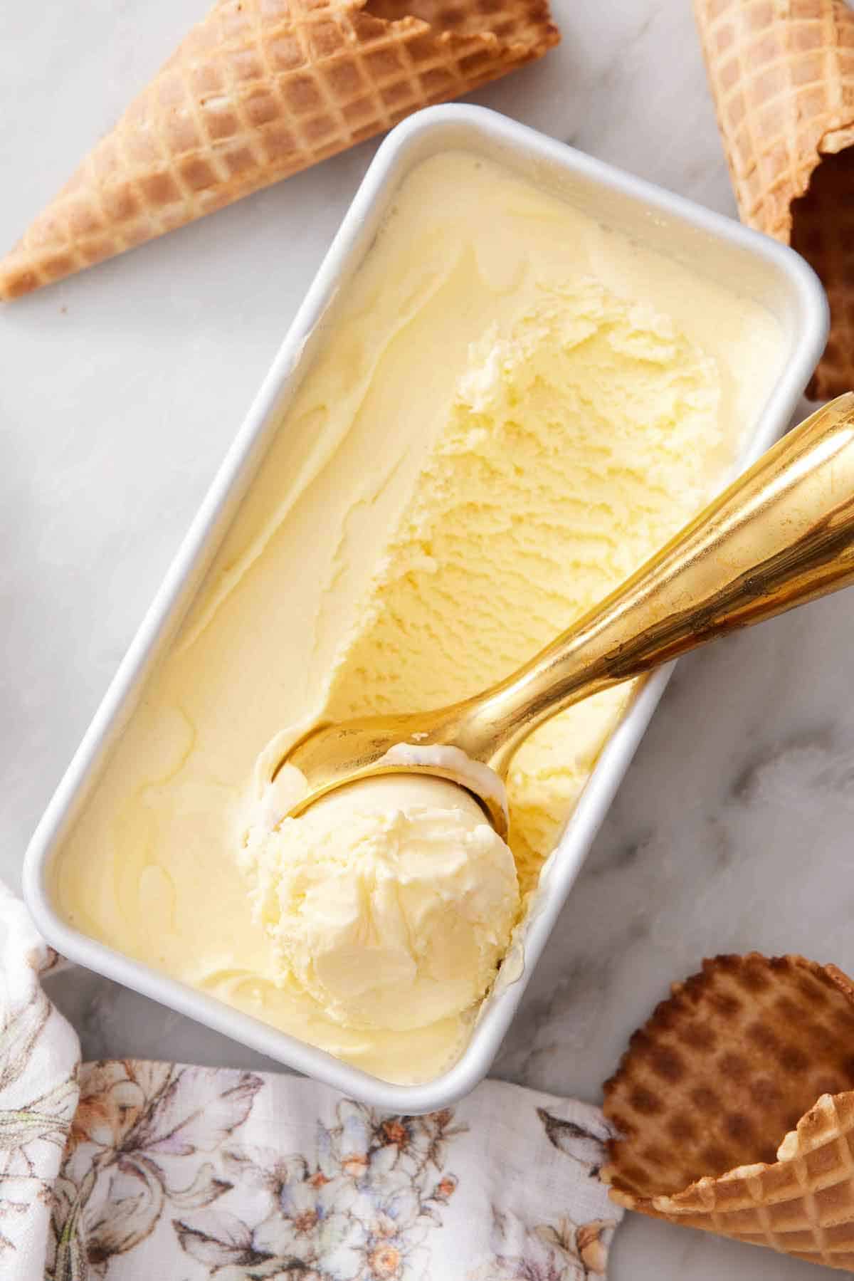 Overhead view of vanilla ice cream in a rectangular baking dish with a scoop run through. Ice cream cones scattered around.
