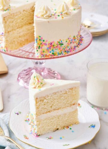 A slice of white cake on a plate with the rest of the cake in the background on a cake stand.