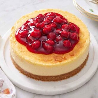 An air fryer cheesecake with cherry sauce on a platter. A cake spatula and stack of plates on the side.