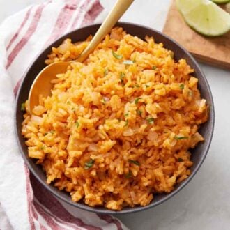 Overhead view of a bowl of Mexican rice with a spoon inside. Cut limes and a napkin on the side.