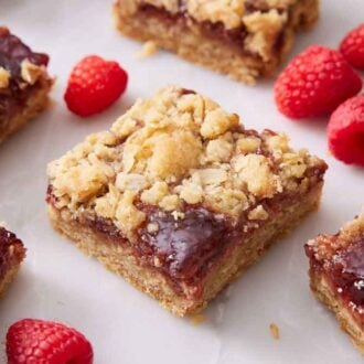 Multiple pieces of raspberry bars on a marble surface with fresh raspberries scattered around.