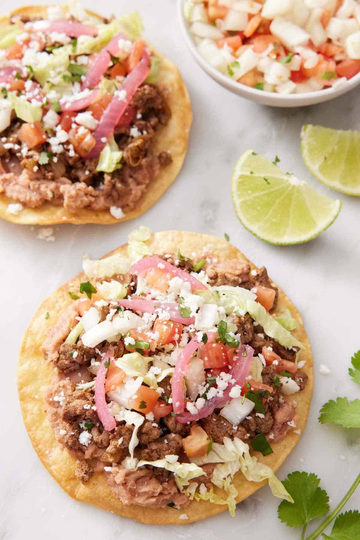 Two tostadas with toppings on a marble surface with two lime wedges and a bowl of pico de gallo.