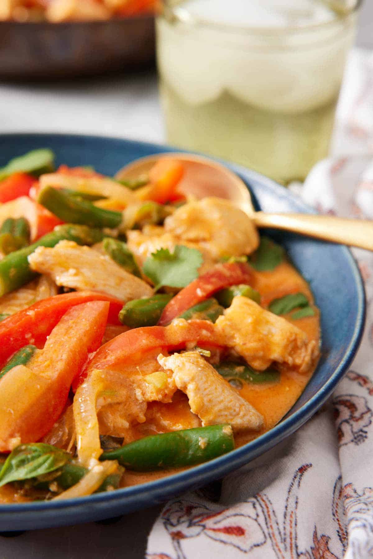 A close up view of a bowl of Thai red curry with a spoon. A drink in the background.