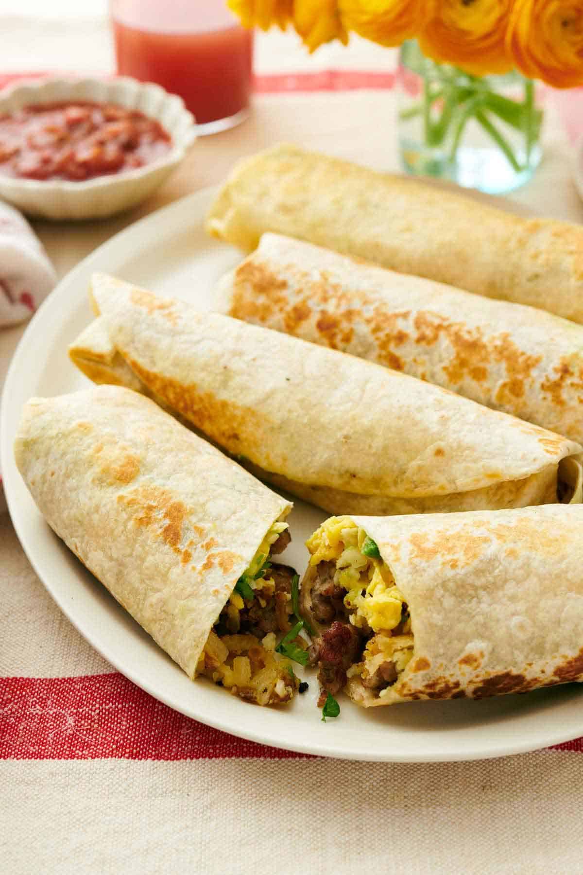 A platter with four breakfast burritos with the one in front cut in half. A bowl of salsa in the background.