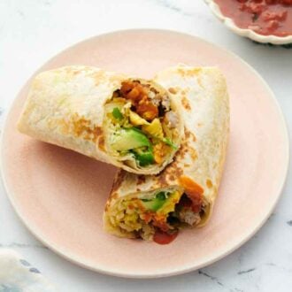 A pink plate with a breakfast burrito, cut in half, with one half leaned on top of the other. A bowl of sour cream and salsa on the side.