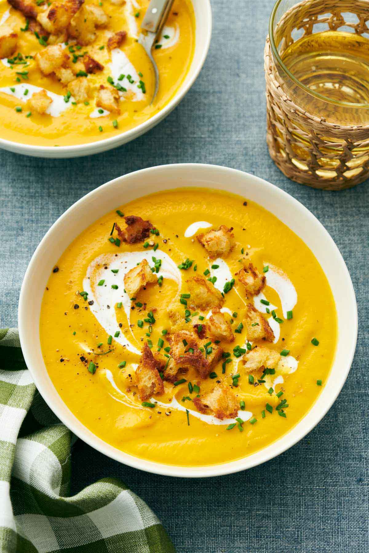 Overhead view of carrot soup in a bowl topped with cream and croutons. A drink and a second bowl off to the side.
