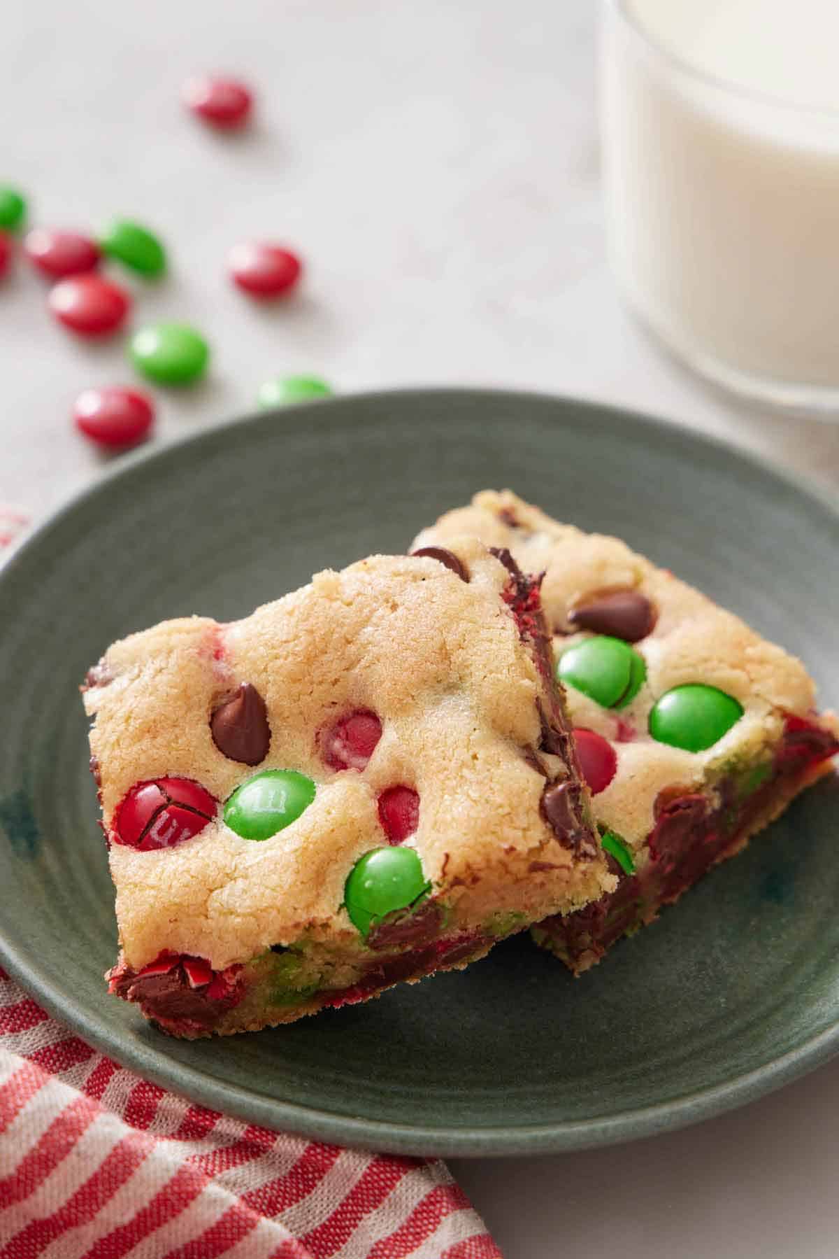 A green plate with two pieces of M&M cookie bars with a glass of milk in the back.