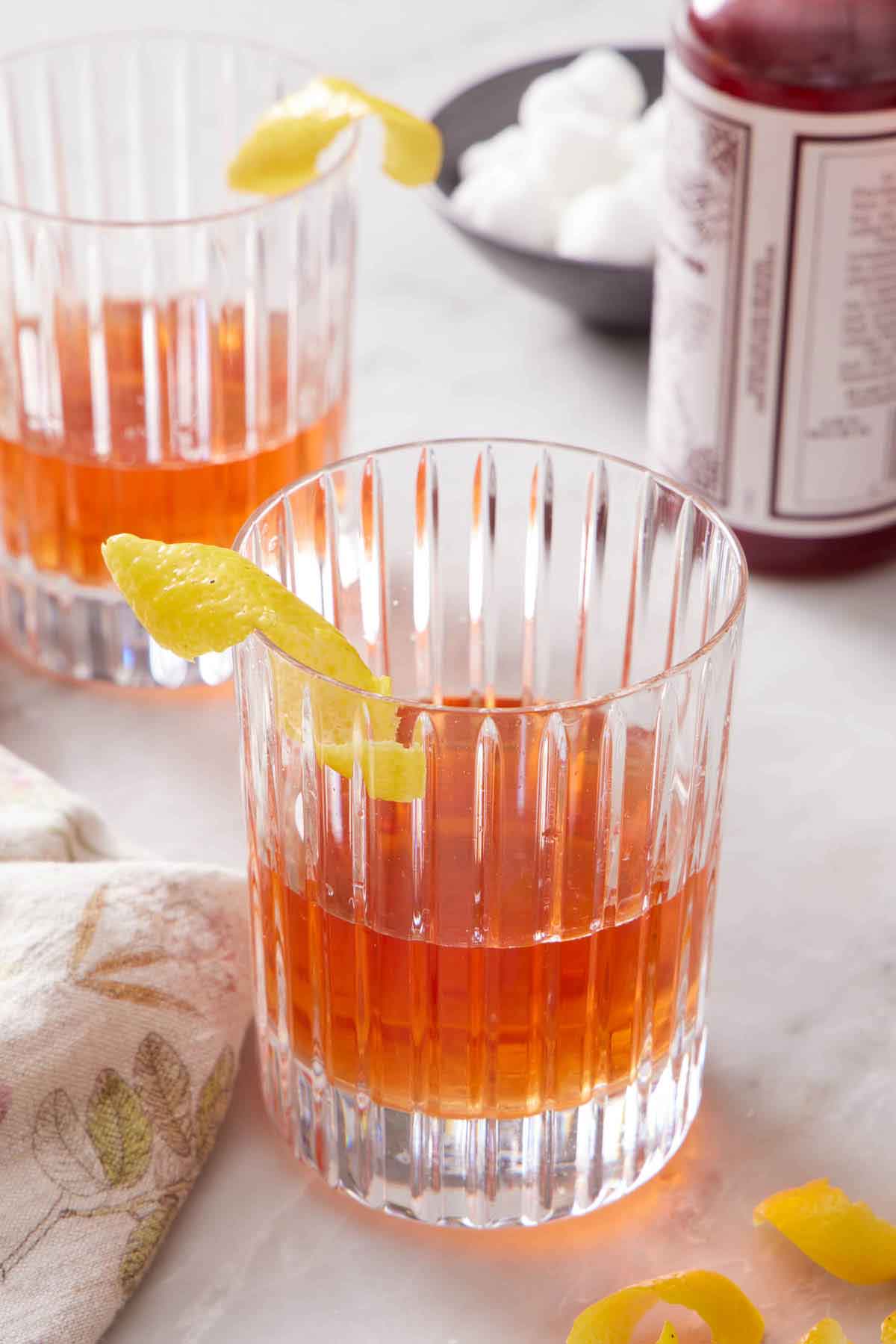 Two glasses of Sazerac with a lemon twist garnish on the rims of the glasses. Sugar cubes and a bottle of bitters in the back.
