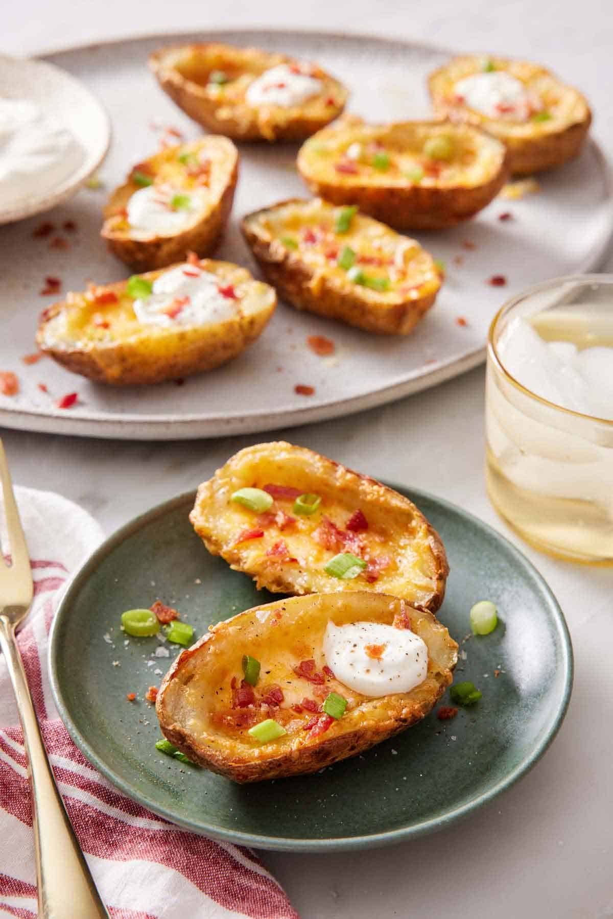 A plate with two air fryer potato skins with a platter in the background with more air fryer potato skins. A drink on the side.